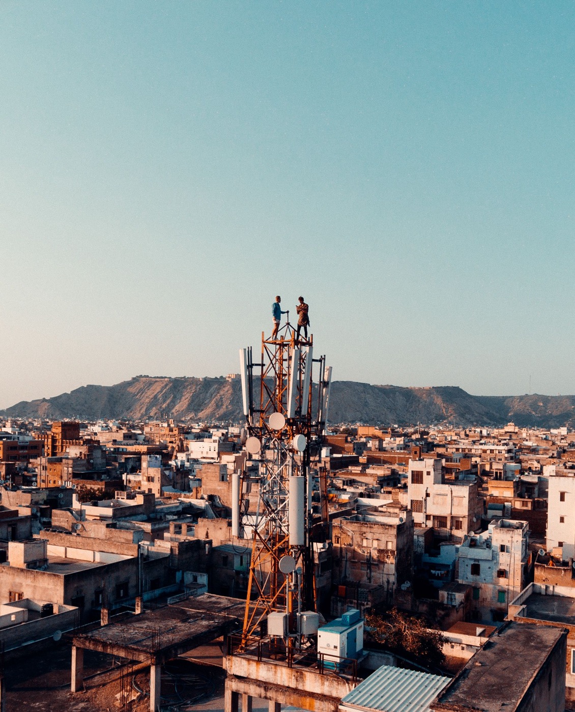 Two technicians stand at the top of a mobile tower above a city.