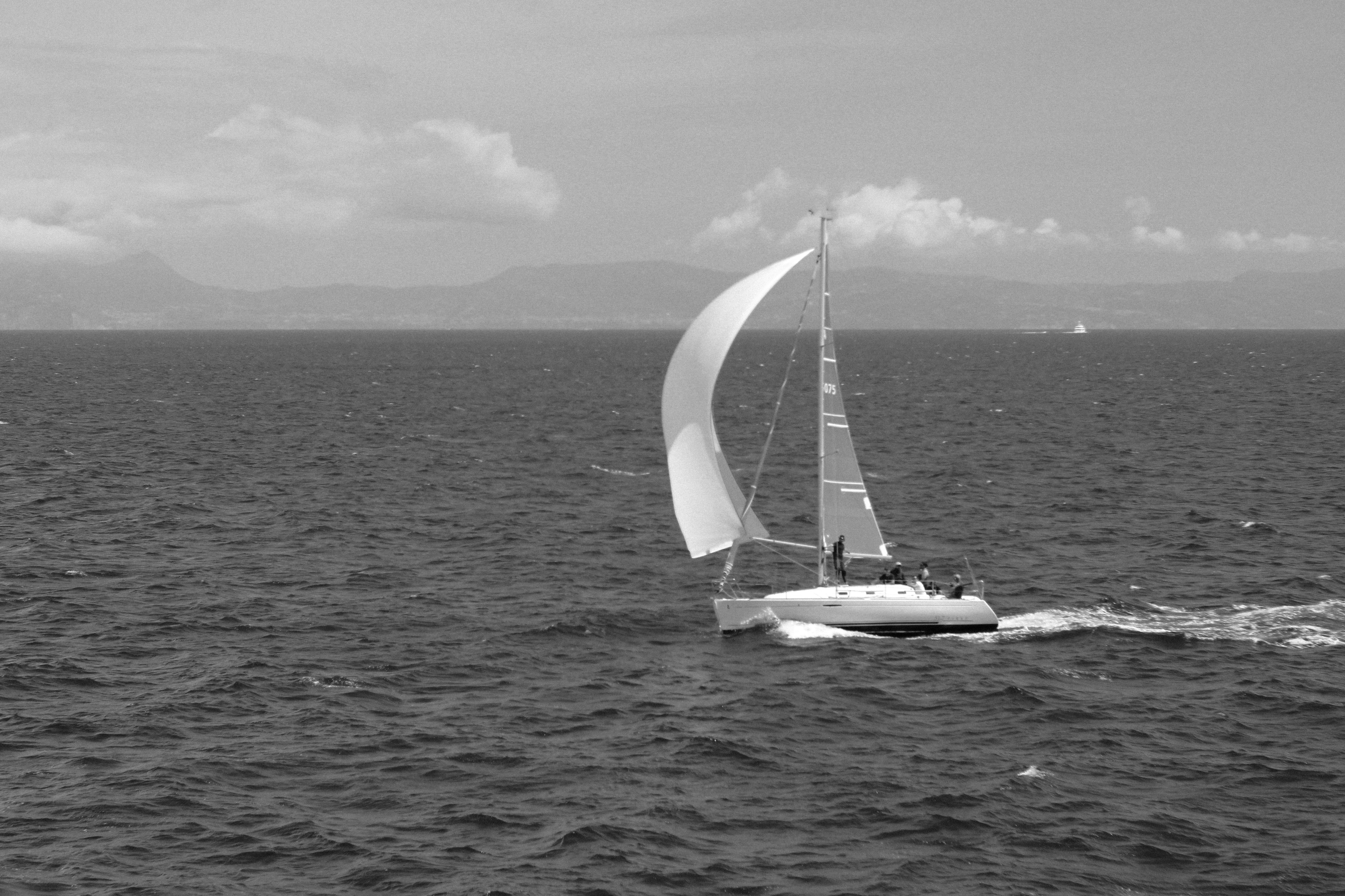 A small sailboat with a full spinnaker crossing open water, a mountain coastline behind it, in black and white.