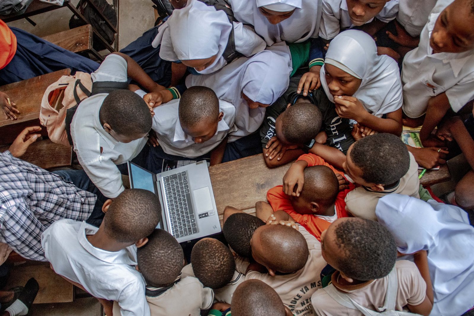 Students in a classroom in Tanzania crowd around a single shared laptop, seen from above.